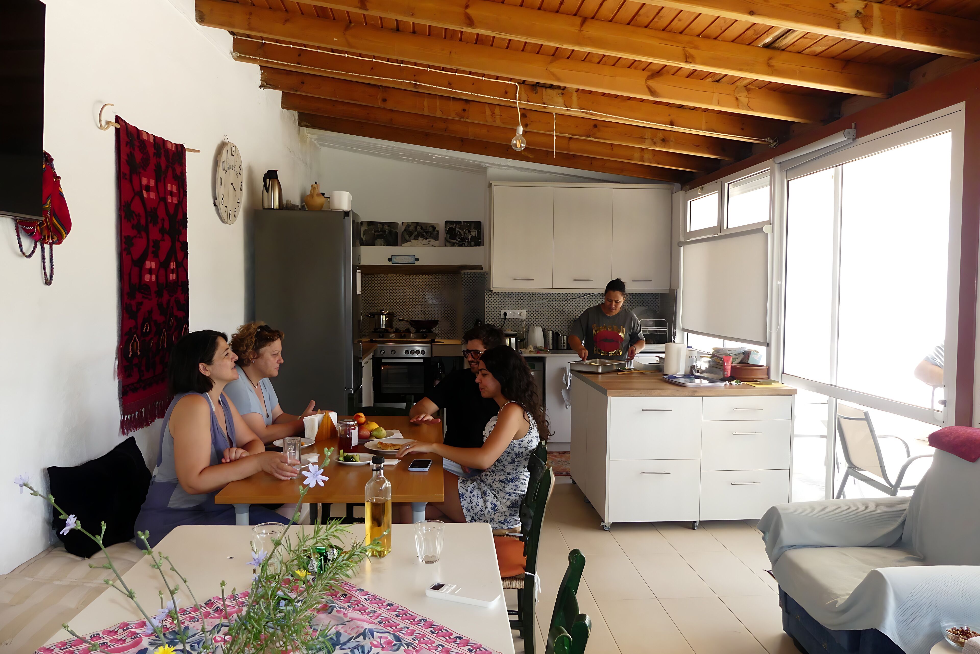 Une cuisine chaleureuse avec des poutres en bois, une table à manger où sont assis des gens, et une vue sur l’extérieur.