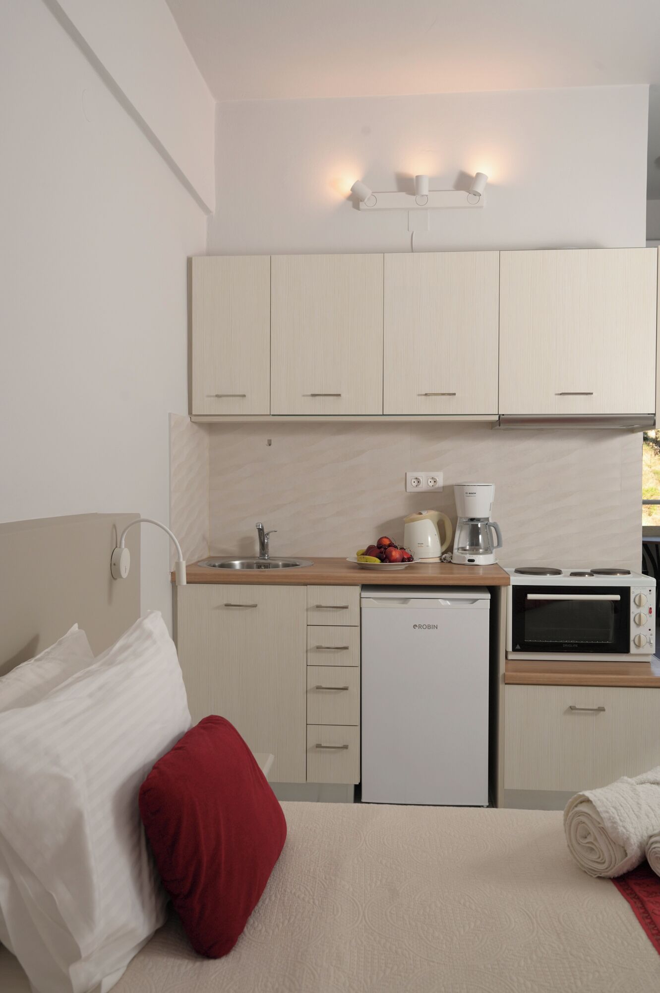 A compact hotel room with a kitchenette, a bed with pillows, and a view of the outdoors.