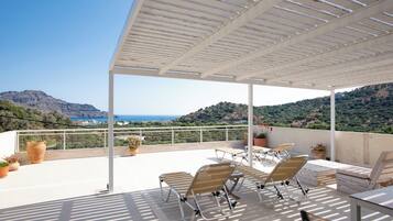 A covered outdoor area with lounge chairs and tables, overlooking a mountainous landscape and the sea.