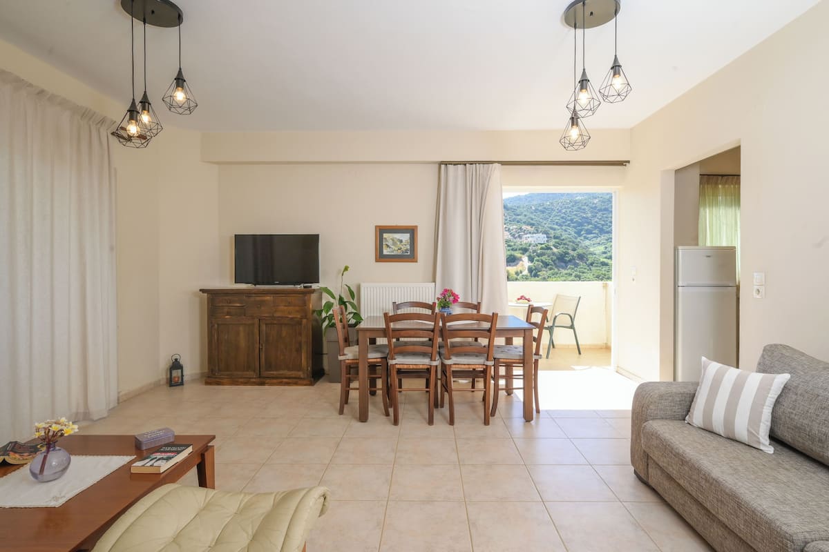 A living room with a sofa, dining table, chairs, a TV, and a view of the outdoors.