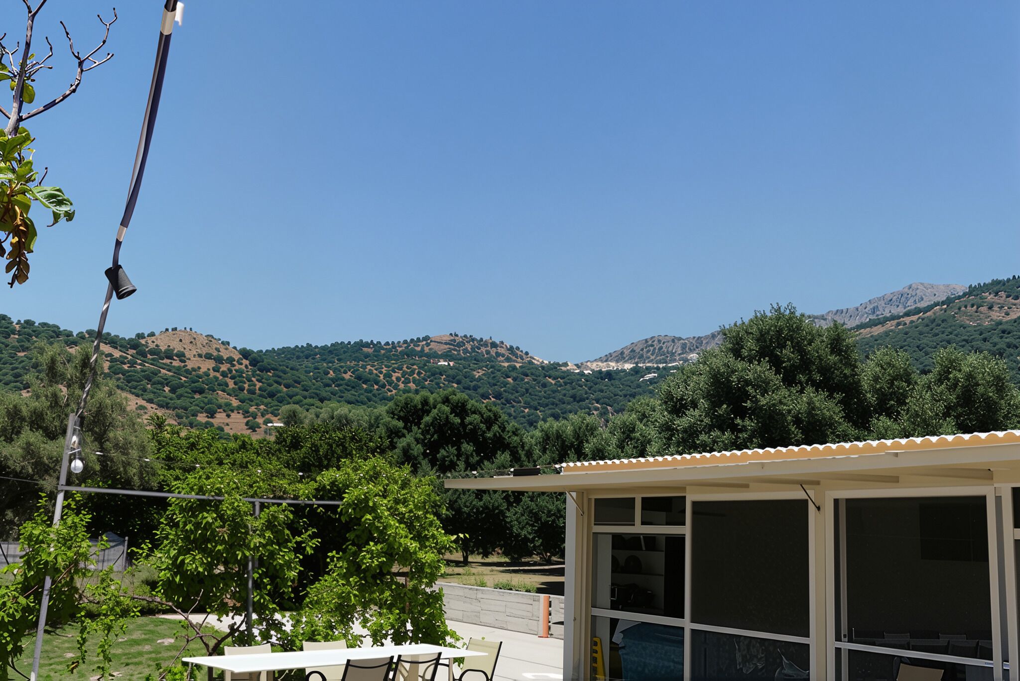 A covered outdoor seating area with a table and chairs, overlooking a hilly landscape with trees and a clear sky.