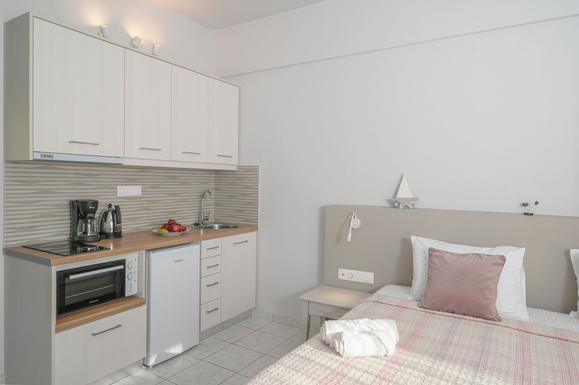 Habitación de hotel compacta con kitchenette, una cama con almohada rosa y una mesita de noche con lámpara.