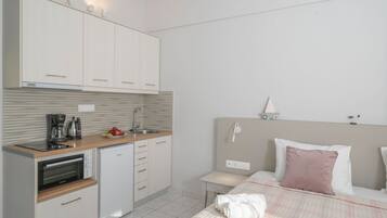Habitación de hotel compacta con kitchenette, una cama con almohada rosa y una mesita de noche con lámpara.