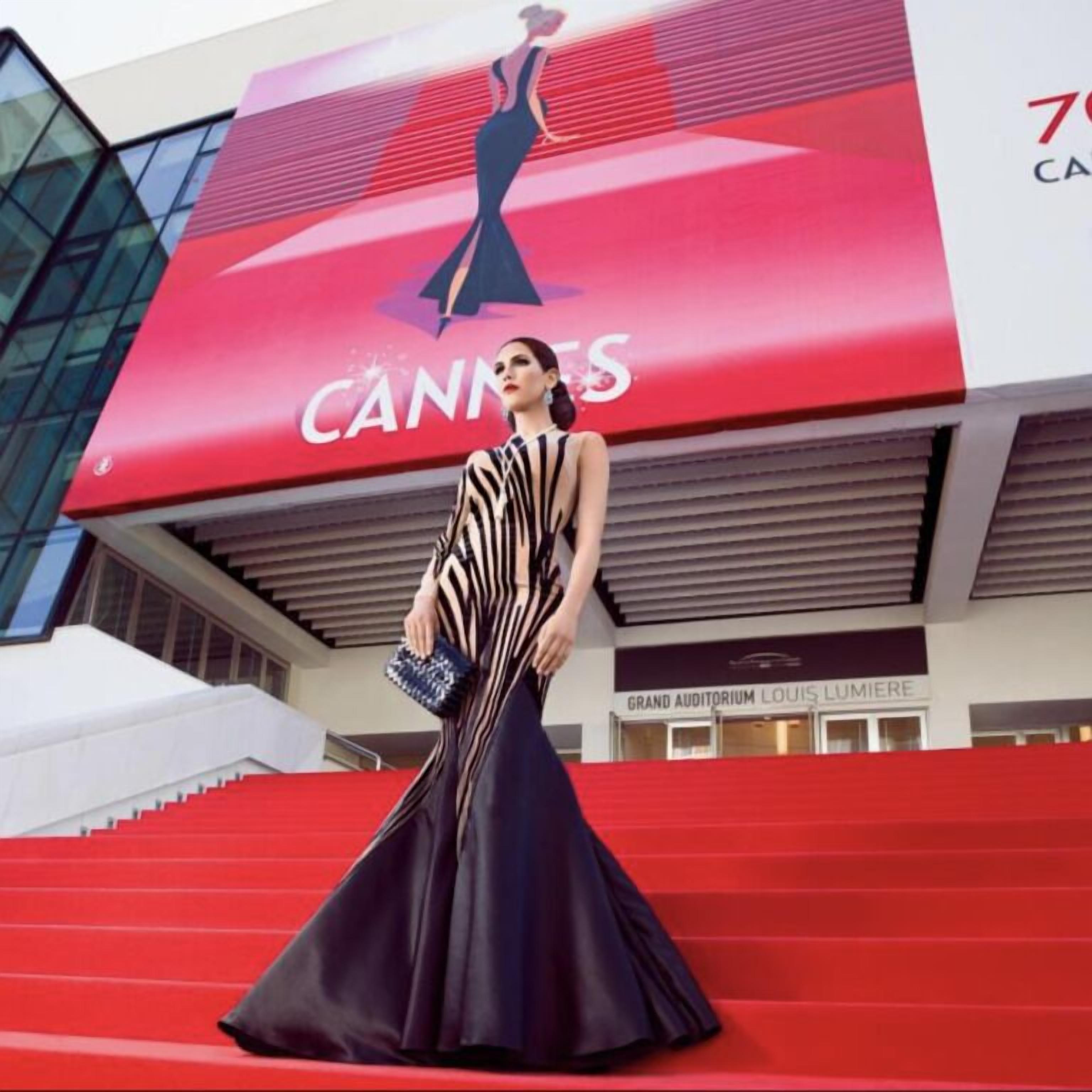 Una persona con un vestido a rayas blancas y negras está de pie sobre escalones rojos frente a un edificio con un gran letrero que dice 'Cannes'.