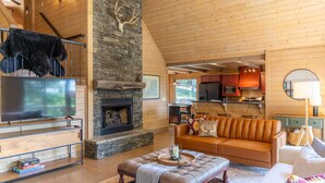 A spacious living room with a stone fireplace, a leather sofa, and a television on a wooden stand. - Queen Ridge Cabin - Great views, secluded, hot tub, fire pit! (West Jefferson)