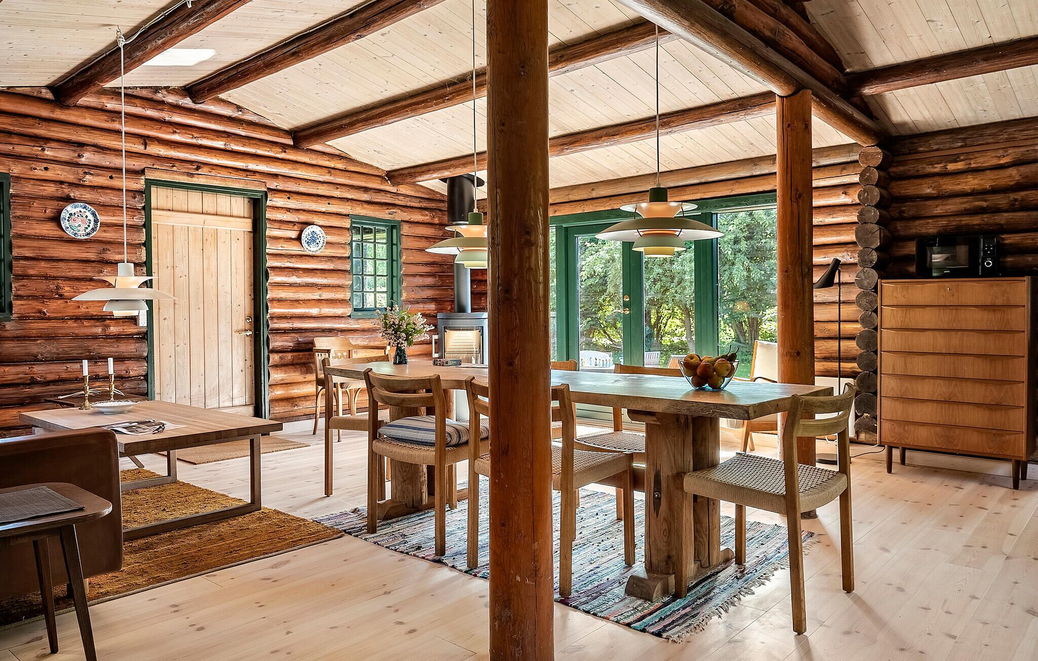 Interior rústico de una cabaña de troncos con comedor, muebles de madera y un amplio ventanal con vistas al exterior.