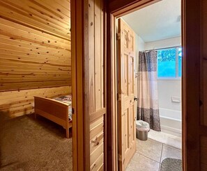 Quarto com teto de ripas de madeira, uma cama, porta para banheiro e janela com vista para árvores.