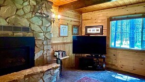 Interior de una cabaña de troncos con chimenea de piedra, televisor de pantalla plana y un amplio ventanal.