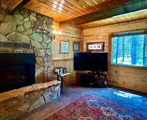 L’intérieur d’une cabane en rondins, avec une cheminée en pierre, un téléviseur à écran plat et une grande fenêtre.