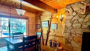 Un acogedor interior de cabaña con una mesa de comedor de madera, sillas, una chimenea de piedra y cuadros en la pared.