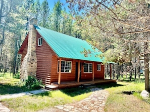 Une cabane en rondins avec un toit végétalisé, une cheminée en pierre et un sentier en pierre menant à l’entrée.