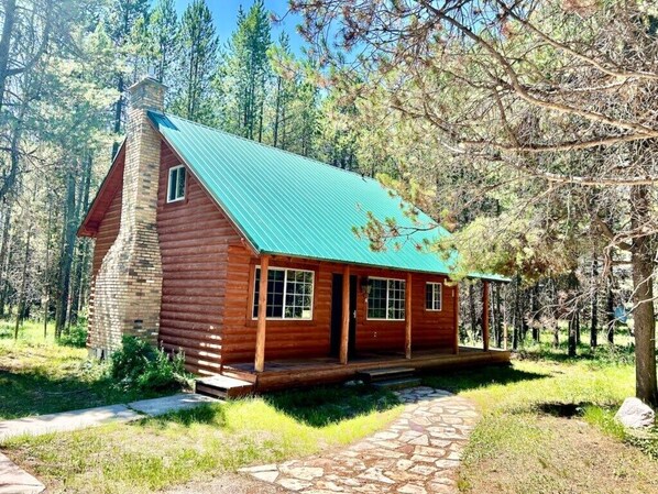 Uma cabana de tronco com telhado verde, rodeada por árvores e um caminho de pedra.