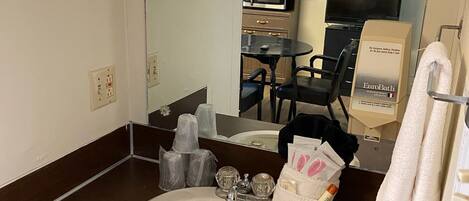 A bathroom with a sink, mirror, toiletries, and a television in the background.