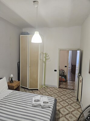 Habitación de hotel con cama, mesita de noche, lámpara, armario, perchero y puerta que da a un pasillo.