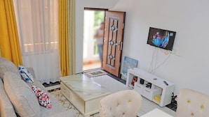 Ein Wohnzimmer mit einer Couch, zwei Sesseln, einem Couchtisch, einem an der Wand befestigten Fernseher und einem weißen Schrank mit Fernbedienung.