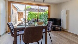 A modern living room with a glass dining table, two chairs, a TV, and a large window overlooking a garden. - Maison Marmillons - Pavilion (AIX LES BAINS)