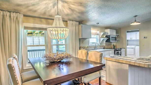A dining area with a wooden table, chairs, and a chandelier. There is a kitchen area with appliances and cabinets visible in the background. - Charming 3-bedroom house in Garden City, featuring two covered porches! (Murrells Inlet)