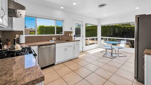 A modern kitchen with white cabinets, a stainless steel range, and a dishwasher. There is a dining area with a table and chairs outside. - Vista Lux Hideaway - A Ryson Property (Palm Springs)