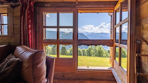 A cozy cabin interior with a leather couch, a large window with a scenic mountain view, and wooden walls.