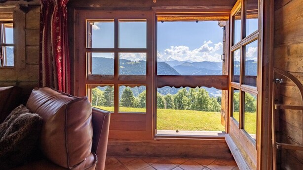 A wooden interior with a leather sofa, large windows offering mountain views, and a rustic ceiling.
