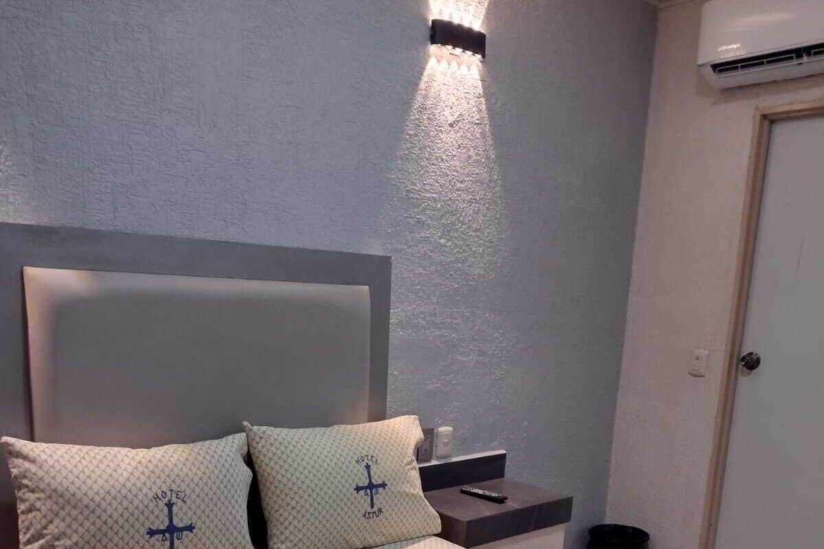 A bedroom with a bed, pillows, a nightstand, a wall-mounted light, and an air conditioning unit.