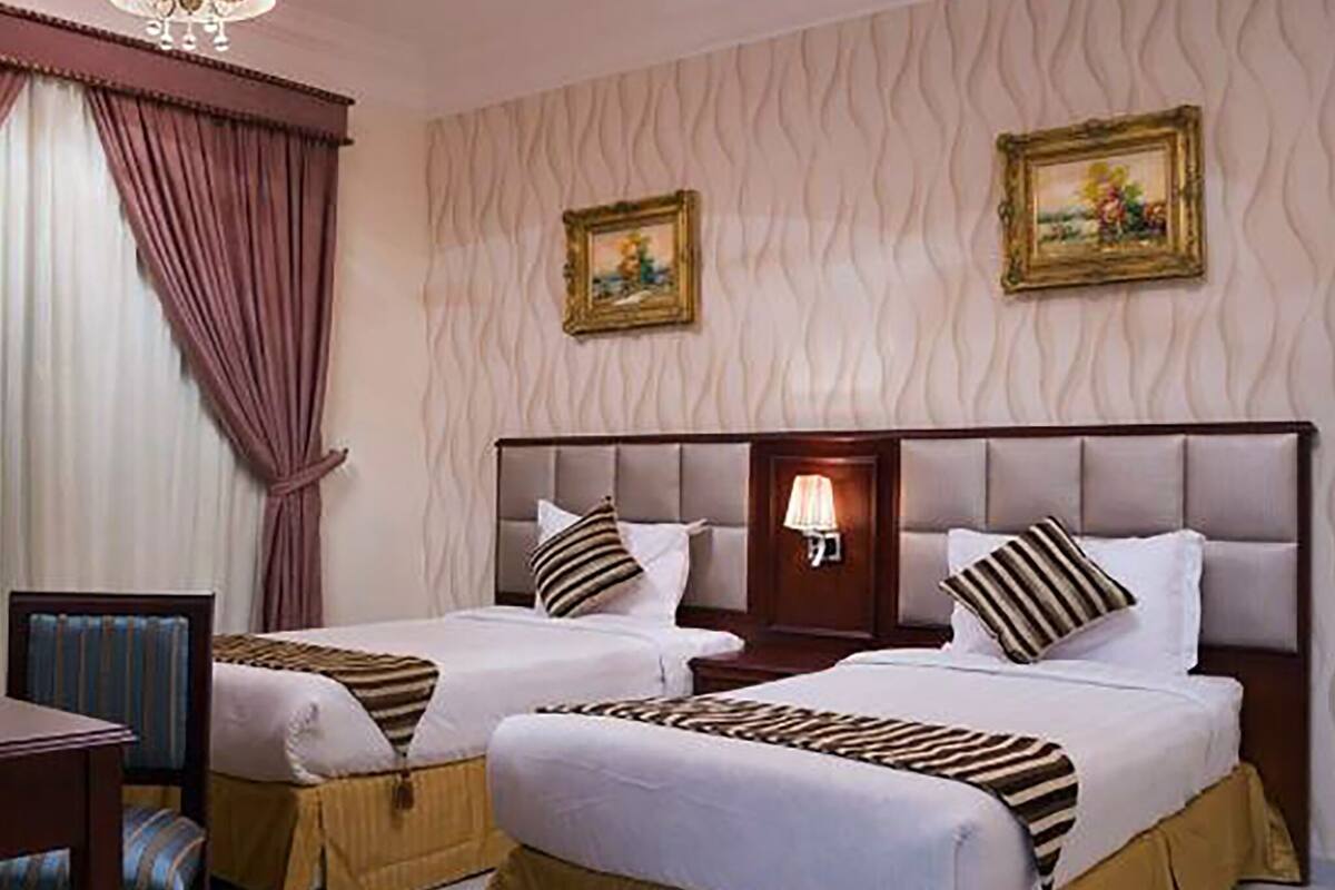 A hotel room with two beds, a nightstand, a chair, and a wall-mounted lamp.