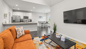 A modern living room with a sofa, coffee table, and a TV. There is a kitchen in the background with white cabinets and appliances. - NEW 3-BR townhome in delightful Spring Hill with WiFi, AC and many amenities (Spring Hill)