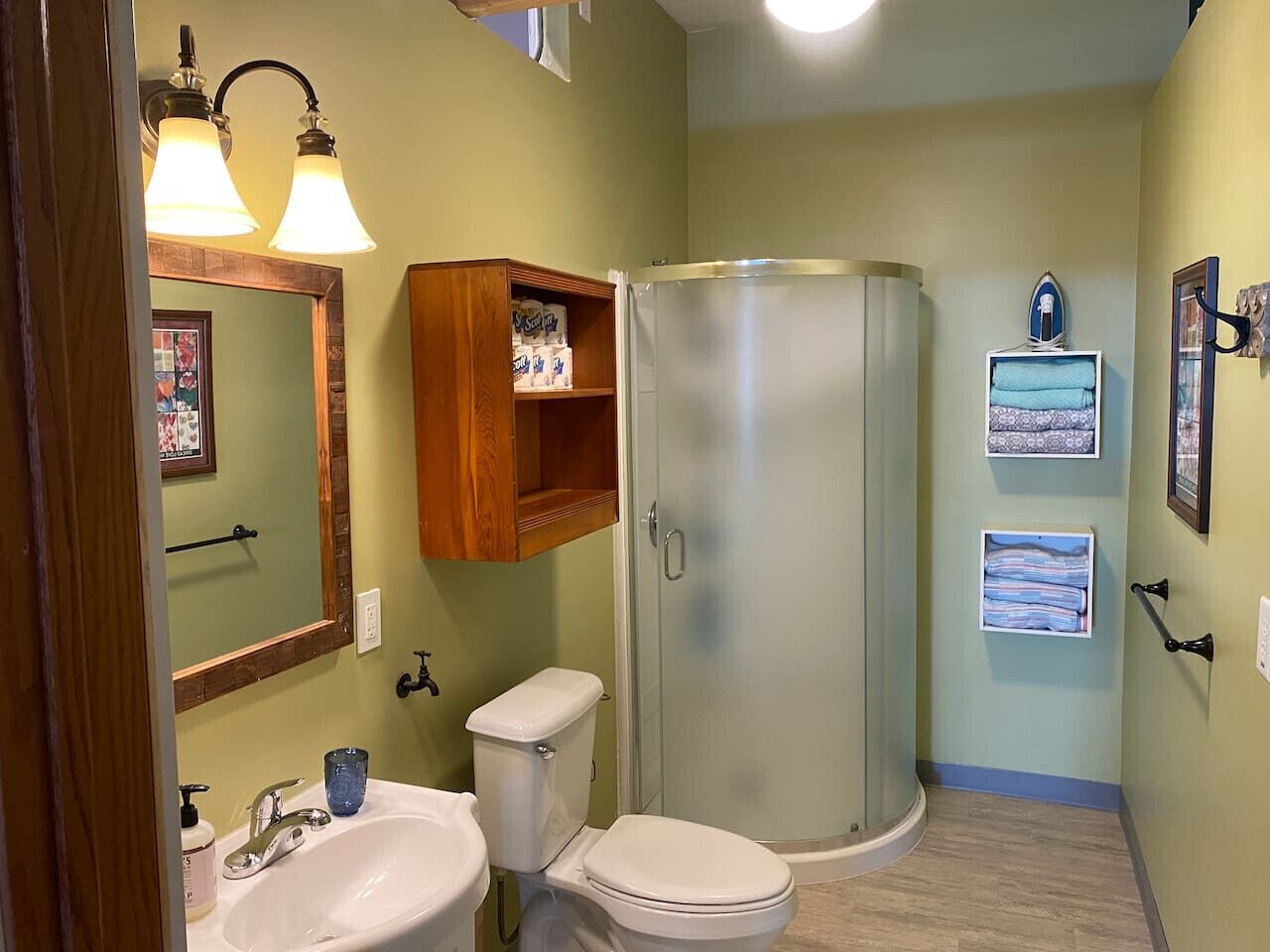 A bathroom with a toilet, sink, and shower. There is a wooden cabinet above the sink and a framed picture on the wall.