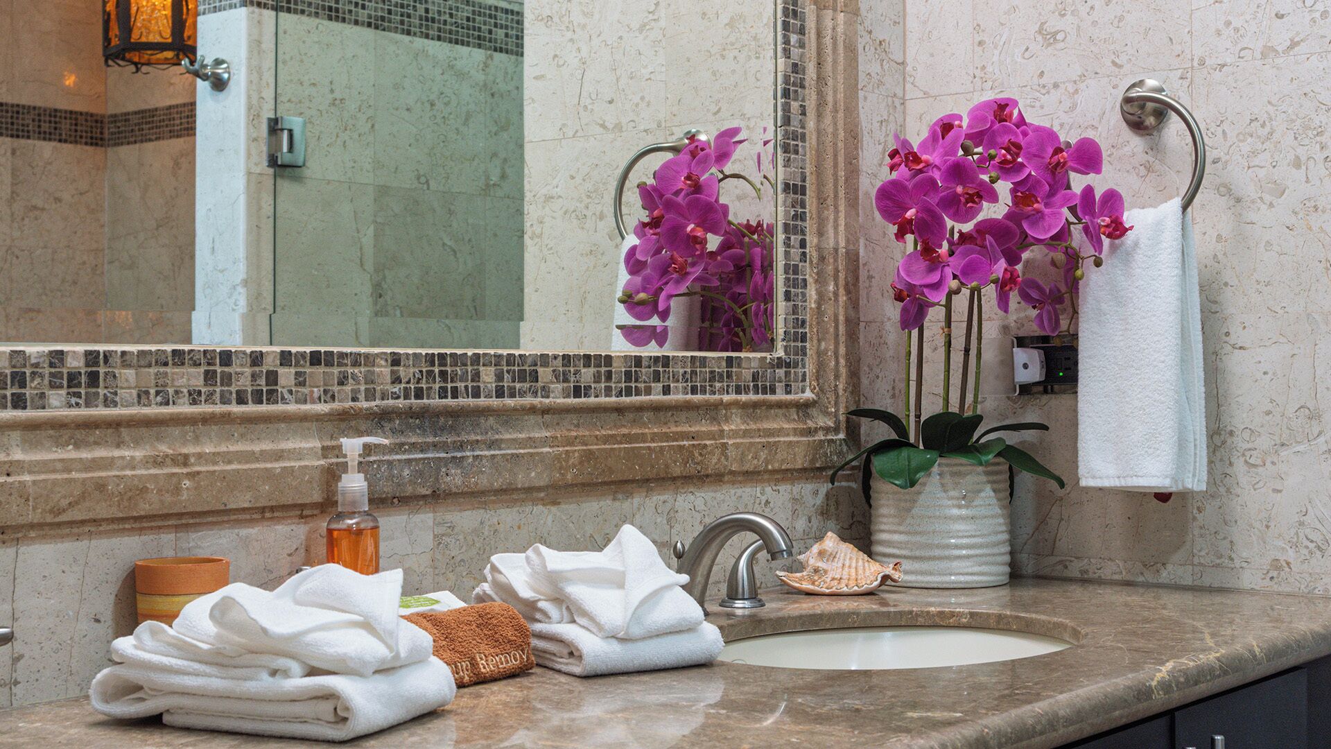 A bathroom with a marble sink, a mirror, a bottle of soap, and a vase with purple orchids.