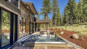 A wooden house with a hot tub on a stone deck, surrounded by trees.