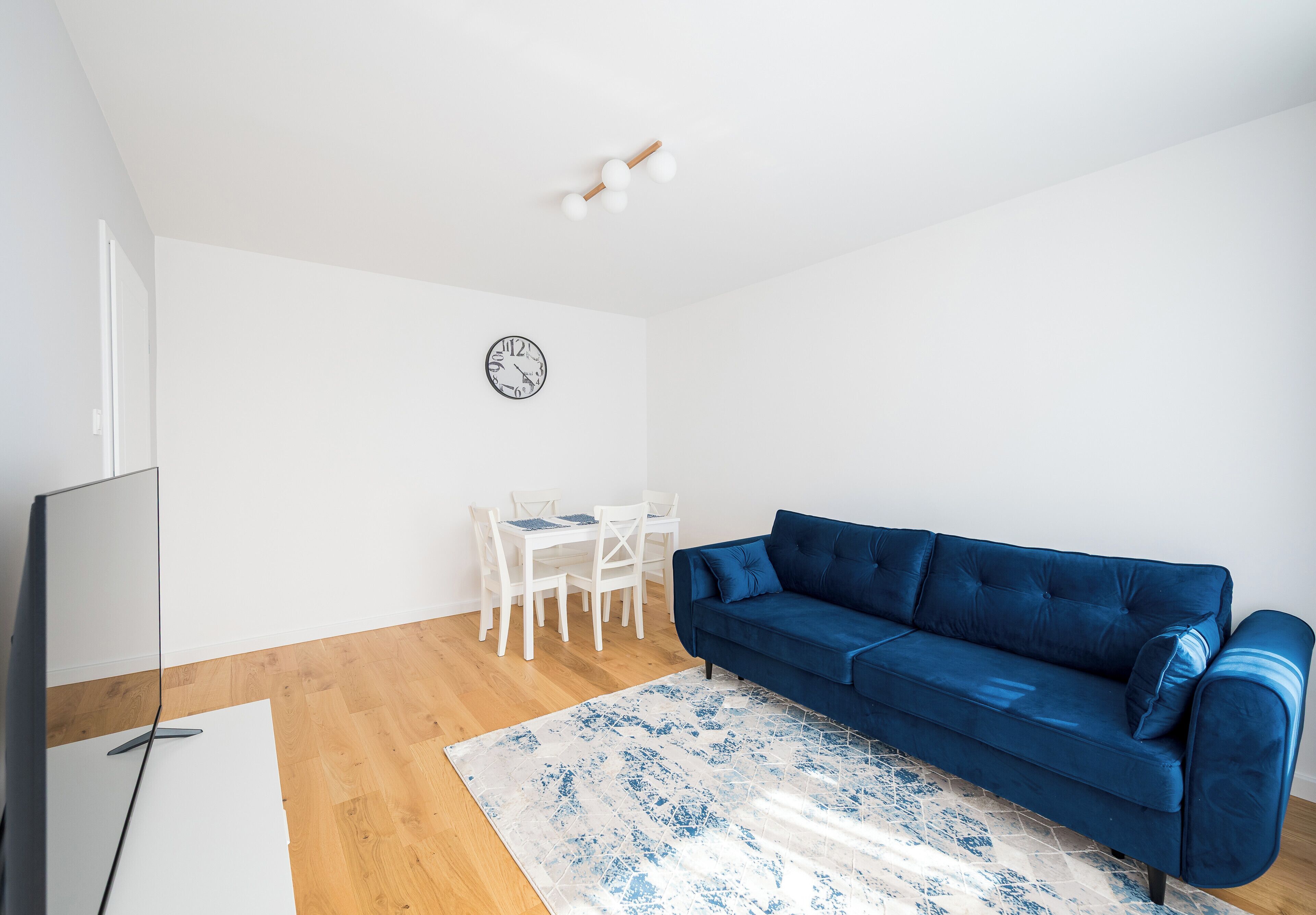 A modern living room with a blue sofa, a white dining table with chairs, a clock on the wall, and a flat-screen TV.