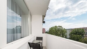 A balcony with a table and chairs, a window with sheer curtains, and a view of buildings and trees. - Stawy Brustmana Homly Apartment (Warszawa)