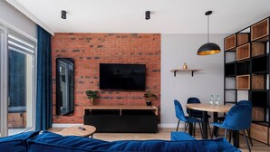 Een moderne woonkamer met een bakstenen muur, een flatscreen televisie, blauwe banken, een houten eettafel met stoelen en een hanglamp.