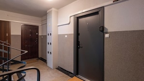 Ein Hotelzimmer mit einer dunklen Tür, einer weißen Wand mit strukturierter Oberfläche, einer Holztür und einem Geländer aus Metall.