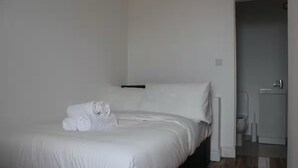 Een net opgemaakt bed met witte lakens en een opgerolde handdoek, een effen witte muur en een zichtbare badkamedeur.