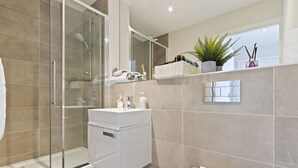 Un baño moderno con cabina de ducha de vidrio, un lavabo blanco con artículos de aseo, inodoro y estante con elementos decorativos y una planta.