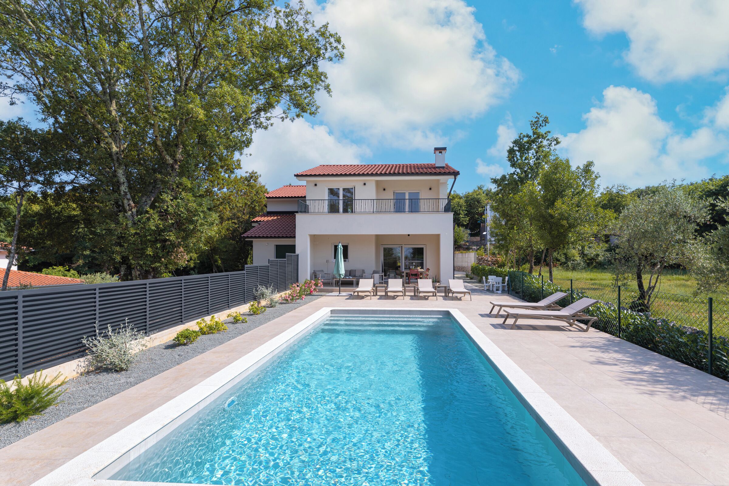 A rectangular swimming pool with a clear blue water surface, surrounded by a tiled deck. There are lounge chairs and a table with chairs on the deck, and a building with a balcony in the background.