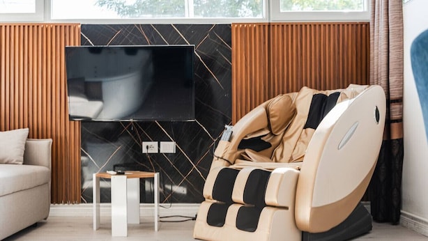 A modern living room with a beige sofa, a massage chair, a small table, and a TV mounted on a black wall. - Green Luxe Suite 2 Private Hot Tub (Shkodër)