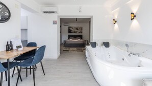 A modern room with a wooden table, blue chairs, a large white bathtub, and a bed with a painting. - Green Luxe Suite 2 Private Hot Tub (Shkodër)