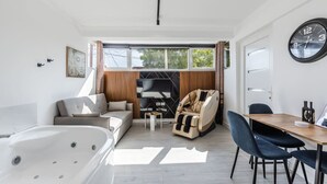 A modern living room with a sofa, a massage chair, a dining table with chairs, a large window, and a clock on the wall. - Green Luxe Suite 2 Private Hot Tub (Shkodër)