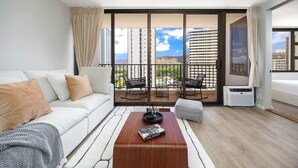 A modern hotel room with a white sofa, a wooden coffee table, a balcony with outdoor seating, and a large window offering a city view.