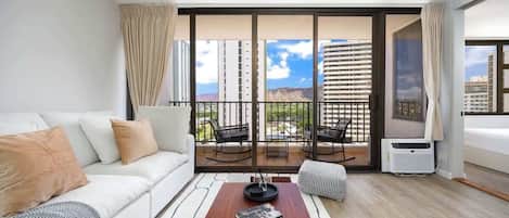 Una habitación de hotel moderna con un sofá blanco, una mesa de centro de madera, un balcón con asientos exteriores y un gran ventanal con vistas a la ciudad.