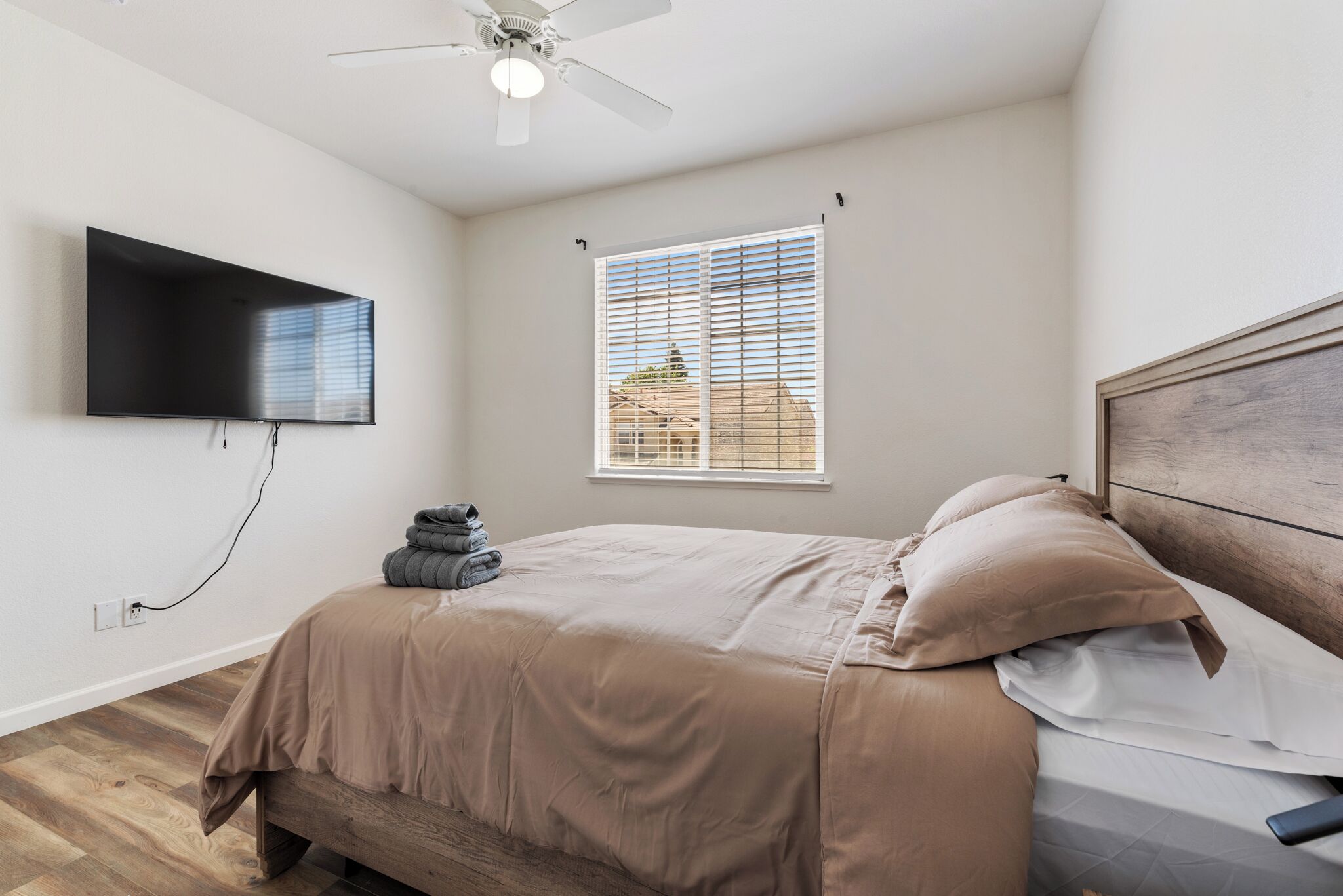 Une chambre à coucher avec un lit, un ventilateur de plafond, un téléviseur fixé au mur et une fenêtre avec des stores.