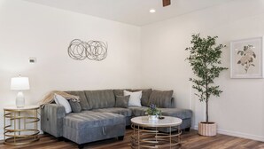 Un salon moderne avec un canapé d’angle gris, une table basse ronde, un lampadaire, une plante en pot et une œuvre d’art murale.
