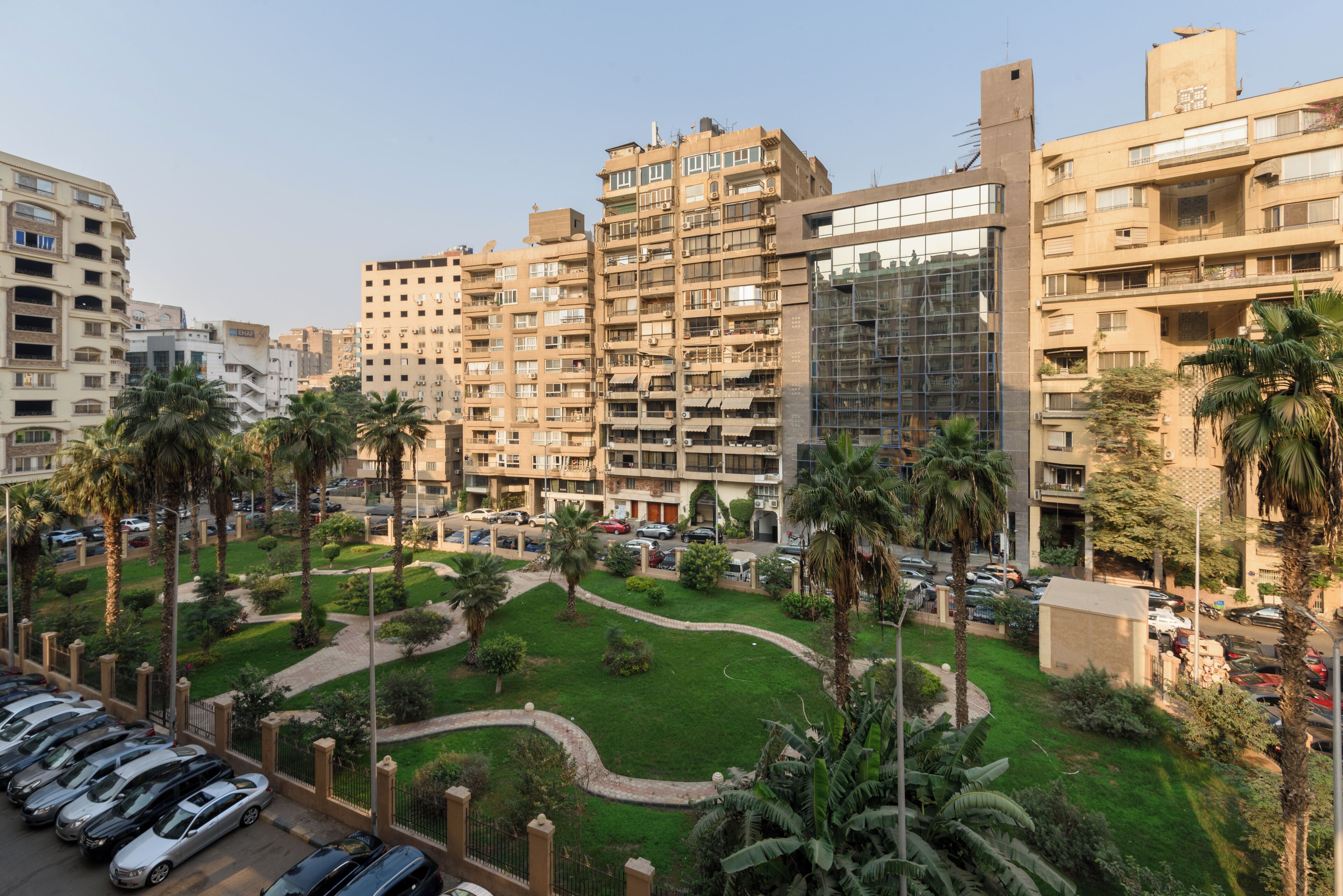A high-rise residential building with multiple balconies, a parking lot, and a well-maintained lawn with palm trees.