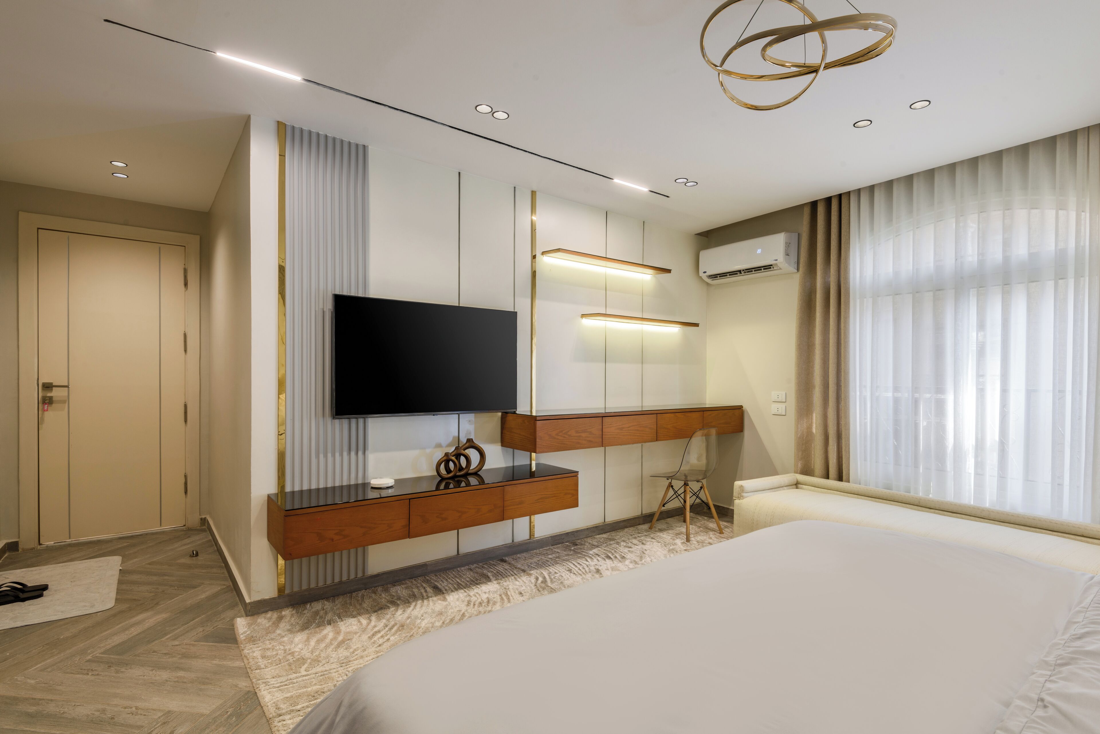 A modern hotel room with a large bed, a flat-screen TV mounted on a wooden console, a small desk with a chair, and a large window with sheer curtains.