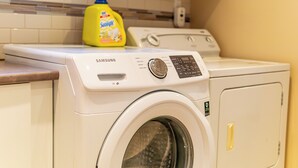 Un lave-linge et un sèche-linge Samsung à chargement frontal, dans une cuisine avec un dosseret carrelé et une bouteille de lessive jaune posée sur le comptoir.