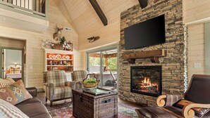 A cozy living room with a stone fireplace, wicker furniture, and a television mounted on the wall. - Huck's Hollow | Hot Tub & Pools (Hollister)