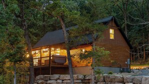 A cabin with a stone wall foundation, wooden exterior, and a balcony with string lights. - Huck's Hollow | Hot Tub & Pools (Hollister)