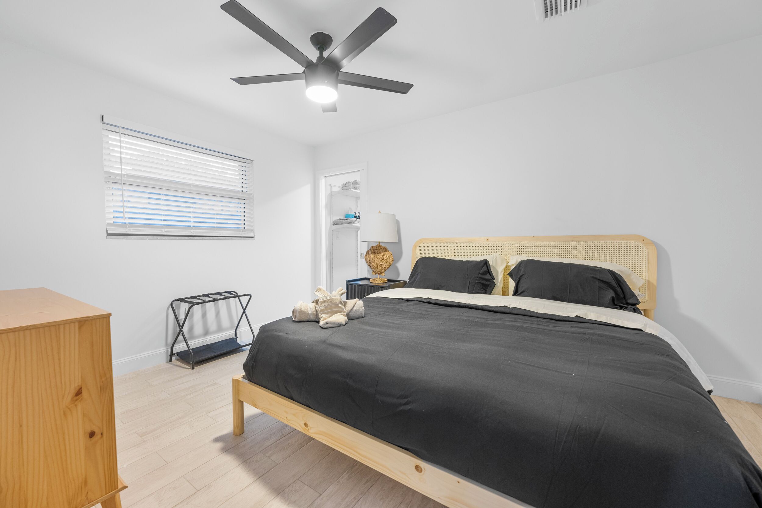 Un dormitorio con cama, ventilador de techo, ventana con persianas, mesita de noche y cómoda de madera.